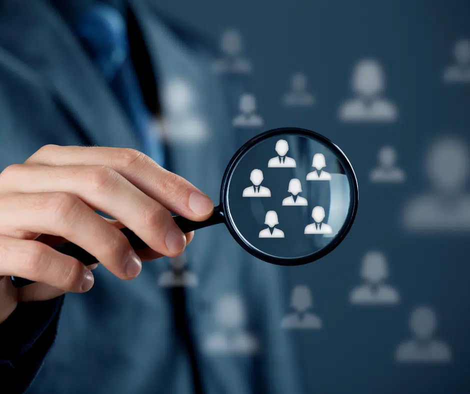 Eine Person hält eine Lupe über gruppierte Personen-Icons, um innerhalb einer Vielzahl unscharfer Symbole eine spezifische Zielgruppe visuell zu identifizieren – Darstellung eines Zielgruppenanalyse-Prozesses.