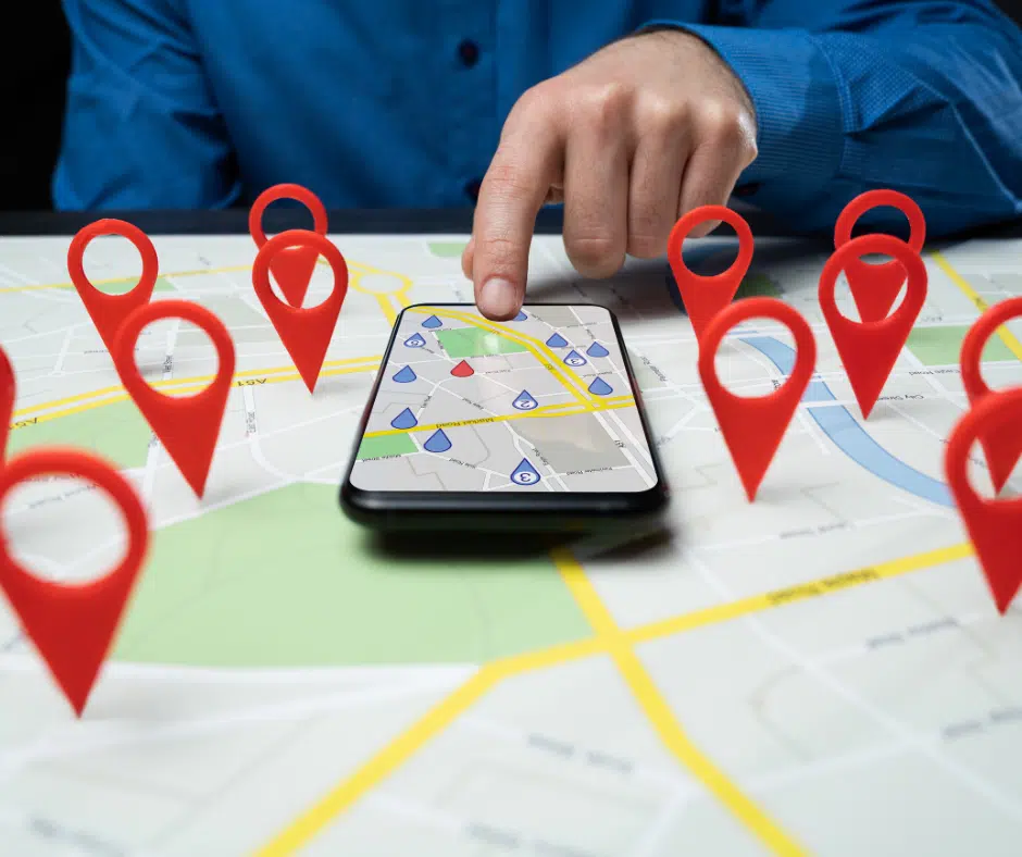Ein Mann zeigt mit dem Finger auf ein Smartphone, das eine digitale Karte mit mehreren Standortmarkierungen anzeigt. Auf dem Tisch liegen zusätzlich viele rote 3D-Standortmarker auf einer gedruckten Karte verteilt.