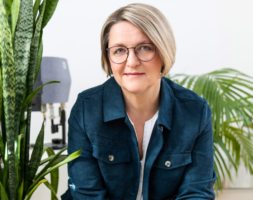 Manuela Kind sitzt vor einem hellen Hintergrund zwischen grünen Pflanzen. Sie trägt eine dunkelblaue Jacke, lächelt in die Kamera und wirkt konzentriert und zugewandt.