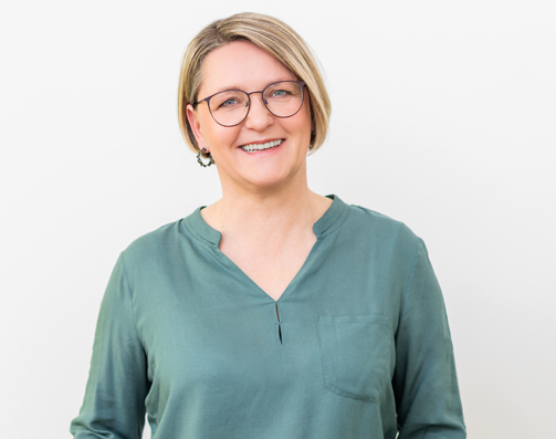 Porträt von Manuela Kind vor hellem Hintergrund. Sie trägt eine grüne Bluse, lächelt freundlich.