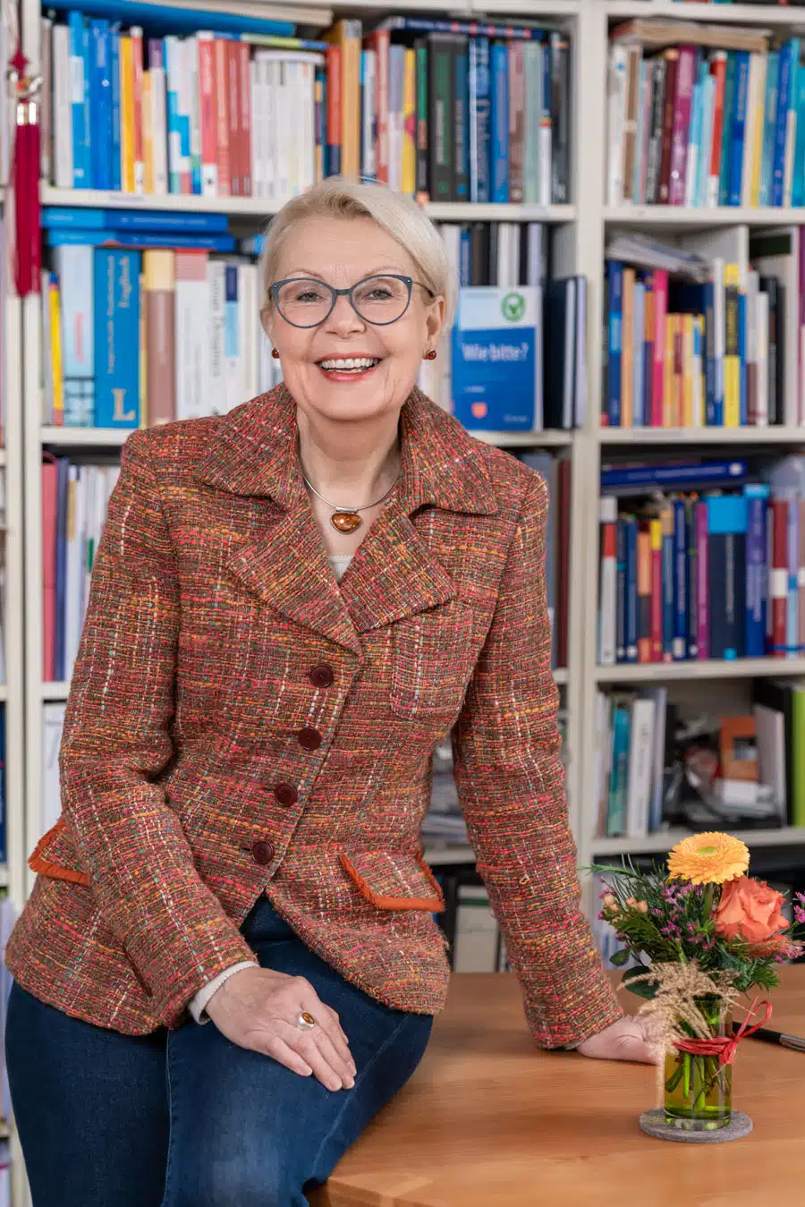 Renate Tewes lehnt lächelnd an ihrem Schreibtisch. Im Hintergrund eine große Bücherwand