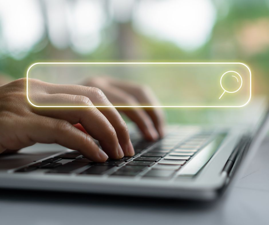 Hände tippen auf einer Laptop-Tastatur, darüber schwebt eine leuchtende Suchleiste mit Lupe-Symbol – Darstellung für Online-Suche oder Informationsrecherche.