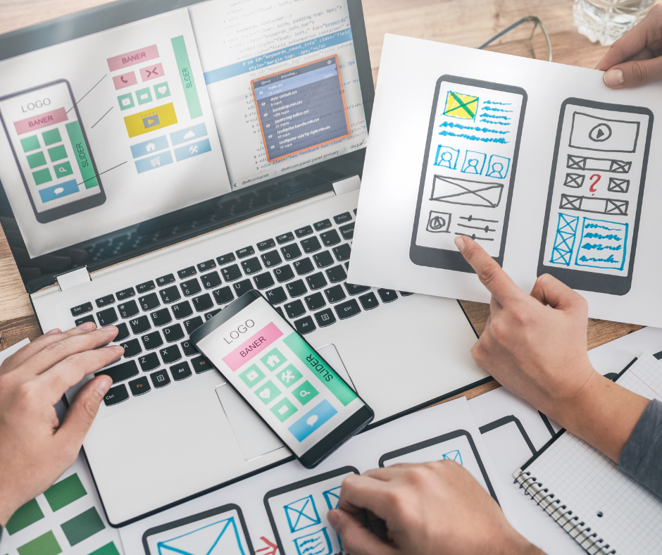 Personen arbeiten an Wireframes und Designkonzepten für eine mobile App oder Website; auf dem Laptop und Papier sind Skizzen von Benutzeroberflächen zu sehen.