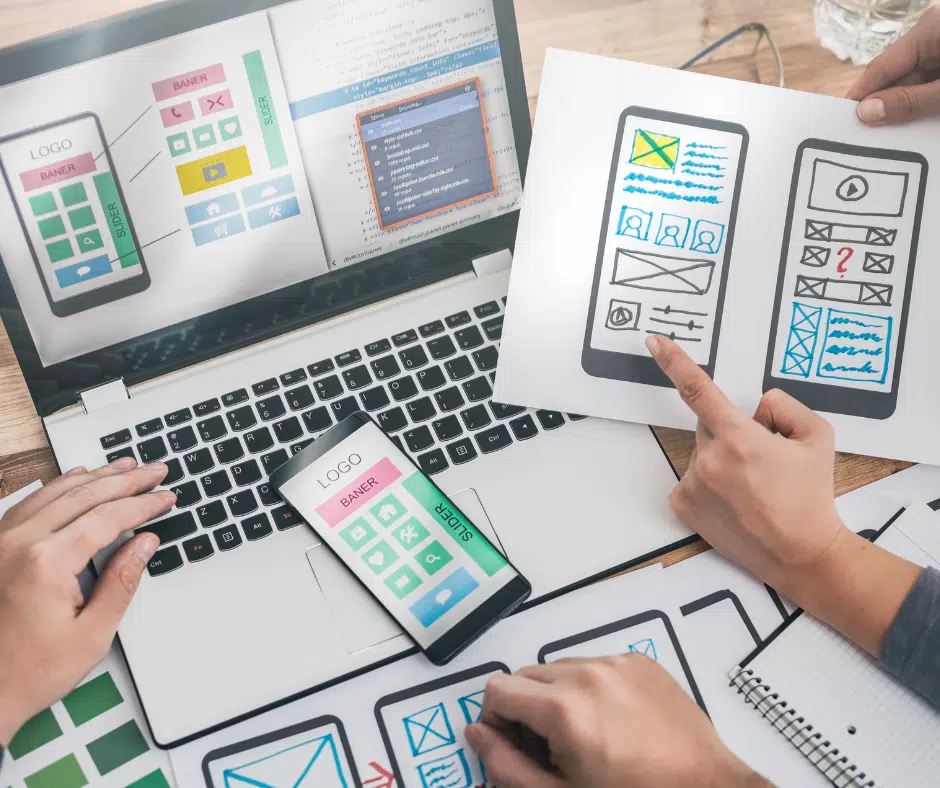Personen arbeiten an Wireframes und Designkonzepten für eine mobile App oder Website; auf dem Laptop und Papier sind Skizzen von Benutzeroberflächen zu sehen.